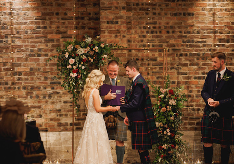 Bride puts grooms ring on his finger whilst celebrant looks on smiling